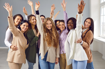 Team of happy diverse business women having fun in the office. Group of cheerful joyful mixed race multiethnic girls standing together, waving their hands, saying hello, smiling and looking at cameraの写真素材