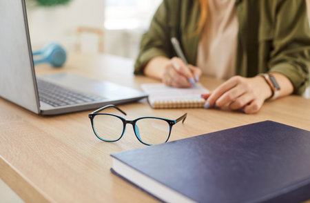 Close-up of eyeglasses on a table with a woman writing in a notepad in the background. Business setting where she is working remotely and engaged in learning in an office environment.の写真素材