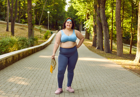 Happy smiling woman in sportswear going to do sport exercises, holding jump rope and looking cheerful at camera standing in summer park outdoors. Workout sport in nature concept.の写真素材