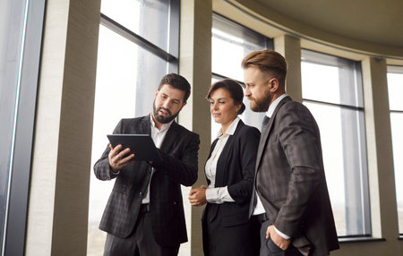 Three business professionals analyzing data on digital tablet in modern office with panoramic...