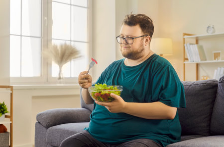 Man eating salad with fork. In a bright living room, the male reflects on healthy nutrition and diet while enjoying fresh greens. Concept: mindful eating and healthy goals.の写真素材