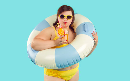 Portrait of funny young woman wearing yellow holiday swimsuit, sunglasses and earrings standing on blue background, holding striped beach ring, and drinking fruit cocktail from glassの写真素材