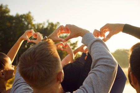 People making heart shape gesture with fingers in sunshine, back view. Crowd of happy young friends showing love and gratitude to nature, greeting summer sun and freedom. Community, hope conceptの写真素材