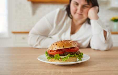 Woman looking at burger lying in front of her, focus on burger. Hungry woman sitting at table in kitchen and eating fast food. Junk unhealthy food addictionの写真素材