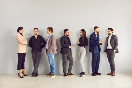 Different groups of business people discuss their work and various interests with each other. Millennial men and women in business casual clothes standing in empty room against gray wall background.の写真素材