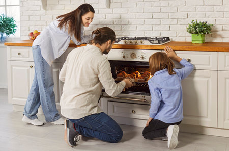 Young father pulling baked muffins from oven standing in modern kitchen at home with son and wife. Cheerful family cooking for dinner tasty dessert together with child boy. Family leisure.の写真素材