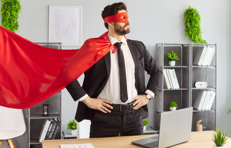 Portrait of confident young funny man in superhero face mask and red cloak standing at the desk on his workplace in modern office and looking proudly to the side. Success and business concept.の写真素材