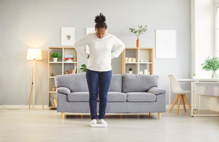 Full length portrait of upset young african american woman wearing casual clothes measuring her weight standing on scales in the living room at home. Diet and slimming concept.の写真素材