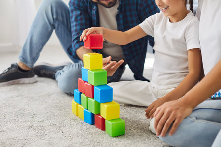Family playing at home, child stacking colorful blocks with help of parents sitting on carpet of living room. Smiling parents and child building balance tower on floor, training coordinationの写真素材