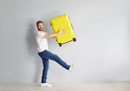 Happy, funny guy carrying suitcase. Full length cheerful, joyful, smiling young man holding yellow suitcase and walking along grey wall background. Traveling, holiday, summer, vacation, trip conceptの写真素材