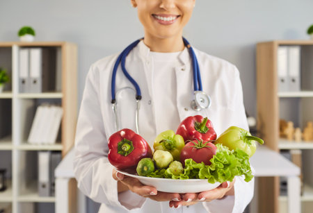 Nutritionist doctor holds vegetables promoting healthy diet. Smiling clinician with stethoscope offers a bowl of fresh salad in medical clinic. Promotes wellness healthcare and nutritionの写真素材