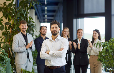 Confident business leader or ceo standing with arms crossed in modern office hallway while smiling professional colleagues applauding in background, celebrating success, achievements and teamwork.の写真素材