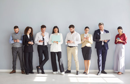 Diverse group of business professionals standing together on a gray background, working online using phones and laptops as a team. Internet technology, and collaboration in an office setting.の写真素材