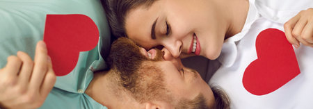 Happy beautiful couple enjoying St Valentines Day. Young man and woman lying together, holding red hearts, touching noses and smiling with eyes closed. Header background, top view from above close upの写真素材