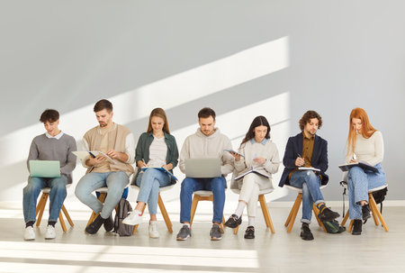 Young people with backpacks, notebooks and laptops sitting in lobby. Male and female fresher or graduate students waiting in line for job interview to use youth internship staff recruiting opportunityの写真素材