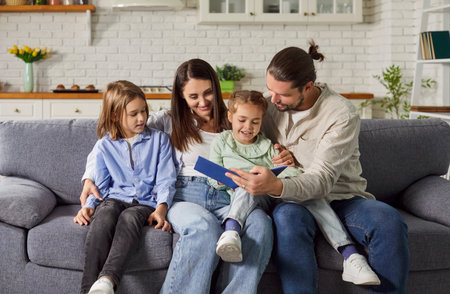 Young happy family reading a book with their little cute daughter and son sitting on sofa at home. Smiling parents spending time with children and smiling. Leisure time together concept.の写真素材