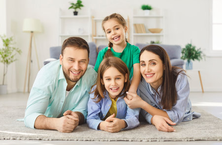 Smiling young happy family of four with two children lying on floor at home and looking cheerful at...