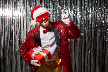 Christmas Santa in sequin costume with gift. Laughing man with beard in red sequined jacket points upward, holding present box before silver tinsel. Festive party joy and new year celebration.の写真素材