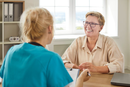 Medical consultation of happy senior woman with doctor in hospital. Smiling patient talking to professional general practitioner, people sitting at desk in clinic for healthcare conversationの写真素材