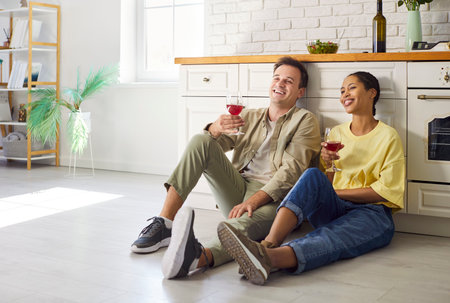 Couple relax at home kitchen, sharing wine. Smiling partners sit on the floor and toast, enjoying love, family time, and bright lifestyle decor nearby. Cozy day together, sharing happiness.の写真素材