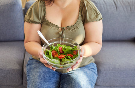 Cropped portrait of a woman wearing casual clothes holding bowl with healthy salad sitting on sofa in living room at home. Nutrition, dieting and weight loss concept.の写真素材