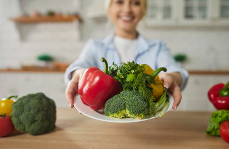 Vegetable plate. Happy smiling woman in blurred background holding plate with broccolis, dill, parsley and peppers over kitchen table. Healthy food, fitness diet concept. Close up, selective focusの写真素材