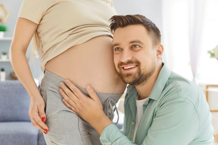 Close up cropped view of happy expectant father cuddling to his pregnant wife belly, listening his baby moving. Loving smiling husband enjoying pregnancy of his wife, excited from future parenthood.の写真素材