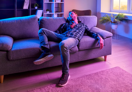 Man on drugs between euphoria and depression. Neon light shows him slumped on a sofa after nightlife, hinting addiction, intoxication, overdose, and loneliness. Despair and mental health crisis.の写真素材