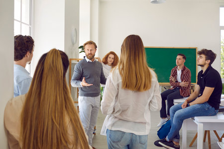 Smart and intelligent male teacher speaks in classroom surrounded by his high school students. Man talks to students at break or informal meeting where they discuss ideas about educational process.の写真素材