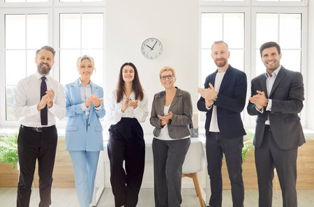 Applauding employees happy clapping hands in office, workers cheering with mentor coach, standing in modern office at corporate meeting, after briefing or workshop, celebrating business achievementの写真素材