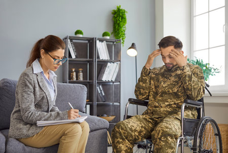 Female psychologist talks to male military veteran soldier wears camouflage uniform sitting in wheelchair during therapy session. Mental health, trauma, PTSD, rehabilitation services support conceptの写真素材