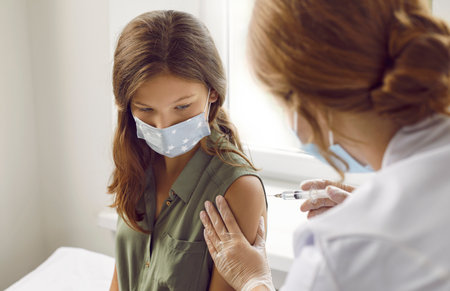 Concept of vaccination against COVID-19 for children and teenagers. Preteen girl receives injection of antiviral vaccine against coronavirus in hospital. Child in mask looks at hands of nurse.の写真素材