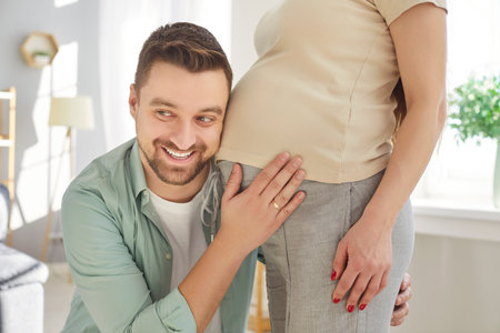 Portrait of happy cheerful handsome man listening to his beautiful pregnant womans tummy and smiling sanding at home. Loving husband enjoying pregnancy of his wife. Couple expecting a baby.の写真素材