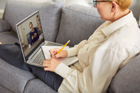 Cropped shot of mature woman sitting on couch, holding pencil and notebook while watching man on laptop screen. Focused lady listening and taking notes during online meeting or educational lecture.の写真素材