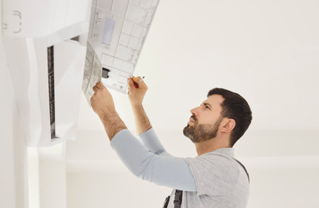 Man working, air conditioner unit mounted on wall, holding instructions, looking intently, focused...