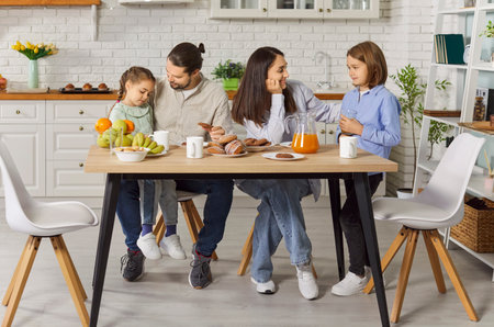 Happy family spending morning together in kitchen, enjoying fresh breakfast with fruits and pastries. Loving parents interacting with their children while eating cookies with fresh orange juice.の写真素材