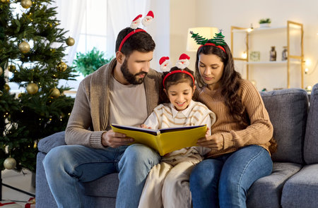 Family reading Christmas book together at home by festive Xmas tree with ornaments. Parents and happy child sitting on cozy living room sofa to read fairy tale story with winter holidays spiritの写真素材