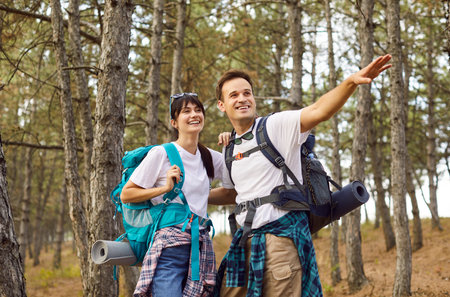 Happy hikers couple with backpacks trekking on forest trail. People walk, carrying mats and gear, enjoying travel and nature. Outdoor exploration, adventure on weekend, and hiking concept.の写真素材
