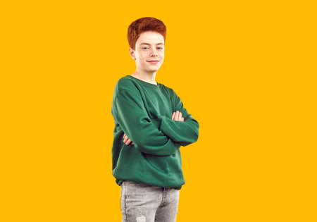 Studio shot of child model in casual clothes. Happy kid posing in fashion studio. Teenage boy wearing dark green sweatshirt standing with his arms folded isolated on yellow orange backgroundの写真素材