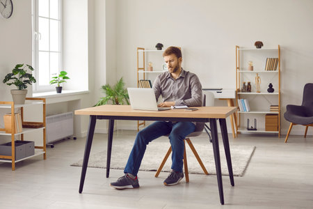 Man working or studying online. Young man sitting at his desk in modern interior at home, using laptop computer, browsing Internet, writing business emails, or studying new educational courseの写真素材