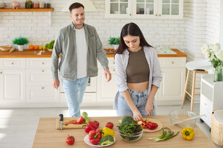 Happy young family couple, woman cooking salad, man helping, healthy vegans at home modern kitchen, vegetables, plate of fresh food at table, health benefits of well-balanced foodの写真素材