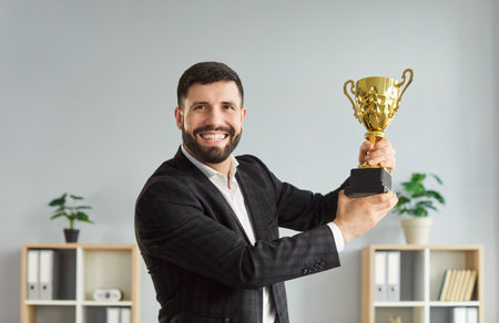 Portrait of happy businessman holding champion cup, celebrating success and achievement in office, focusing on career in a professional setting with a sense of business triumph and joyful recognition.の写真素材