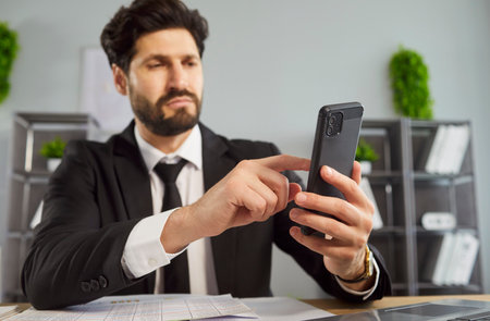 Serious corporate worker in formal wear using mobile phone or chatting with partners, rests after long work with business papers, concentrated in screen. People, technology, communicationの写真素材