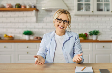 A young woman wearing casual clothes looking at camera having video call sitting at the desk at home. Satisfied confident girl chatting online or having video conference in kitchen.の写真素材