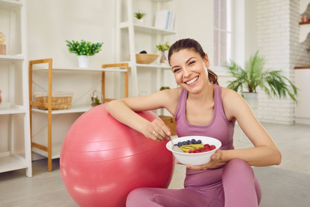 Portrait of a young sporty cheerful slim woman wearing sportswear eating healthy food after working out at home sitting on the floor on yoga mat. Healthy nutrition and lifestyle concept.の写真素材