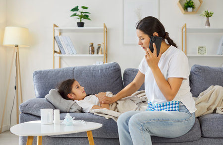 Sick child with fever watching mother. Caring parent calls doctor for treatment advice about virus and cold by smartphone at home. Girl lies under blanket. Parental care during illness.の写真素材