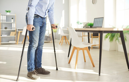 Injured man standing at workplace in office using crutches.Male person with disability wearing...