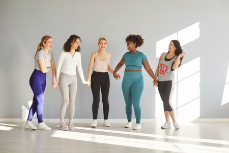 Group of motivated mixed race women dressed in sportswear, holding hands together and looking gladfully at each other, stand in full length against grey background, going in for sport regularlyの写真素材