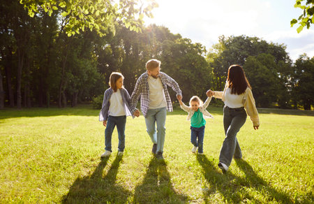 Four family members having fun at park during sunny summer day, walking together and holding hands, having good relationships, enjoying nice day. Parents and children resting outdoorsの写真素材