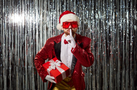 Secret Santa game, man posing playfully in red outfit, hat, jacket, holding gift making shush gesture, party fun, creating playful and secretive atmosphere, surprise gift or hidden festive funの写真素材